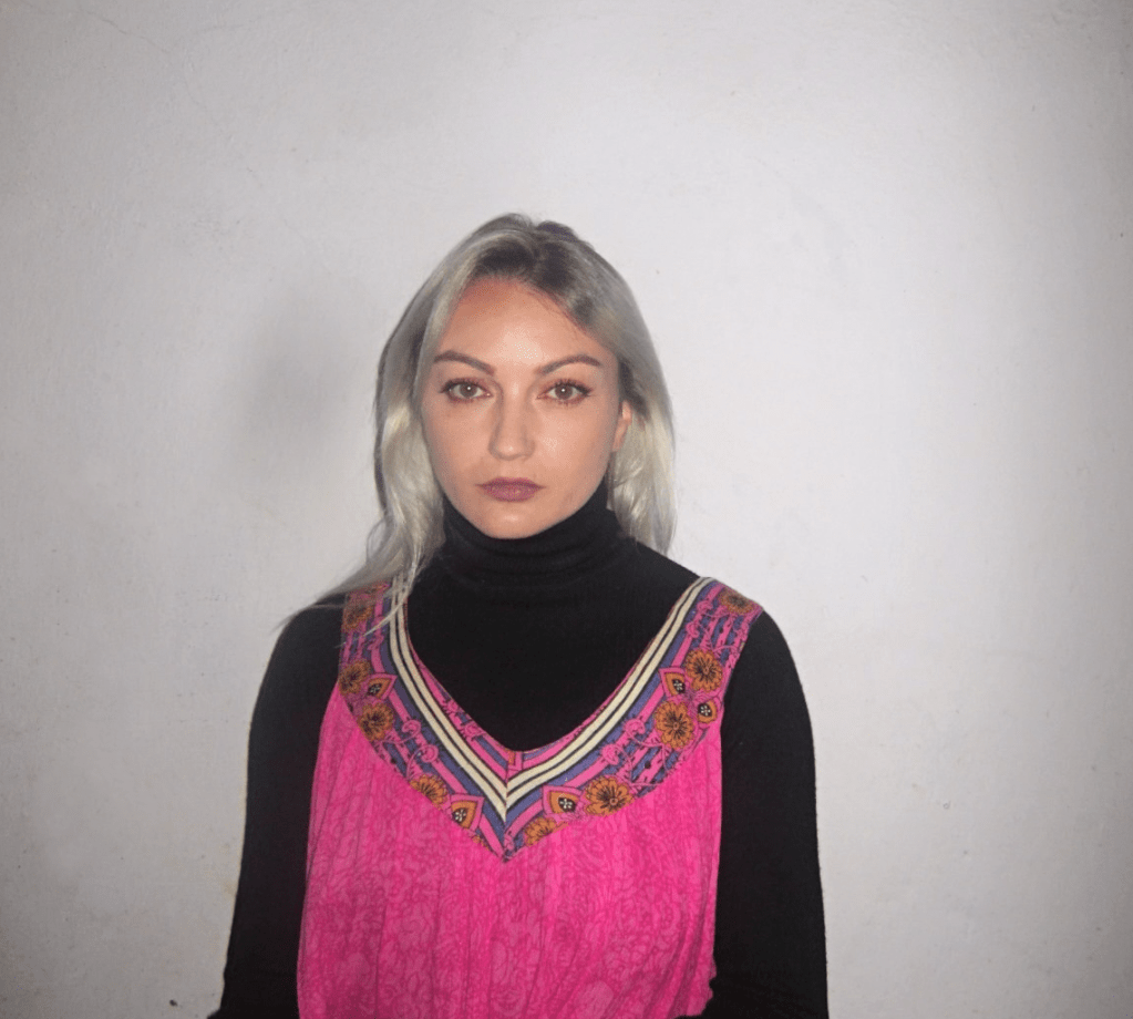 A photo of Casica, artist's name of Casandra Schaut, standing in front of a white wall. She's wearing a pink bohemian dress and black turtleneck sweater underneath.